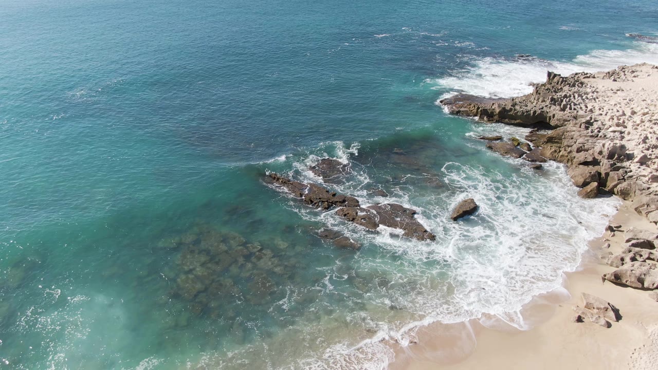 Aerial view of turquoise waves breaking over rocks beside a sandy beach.