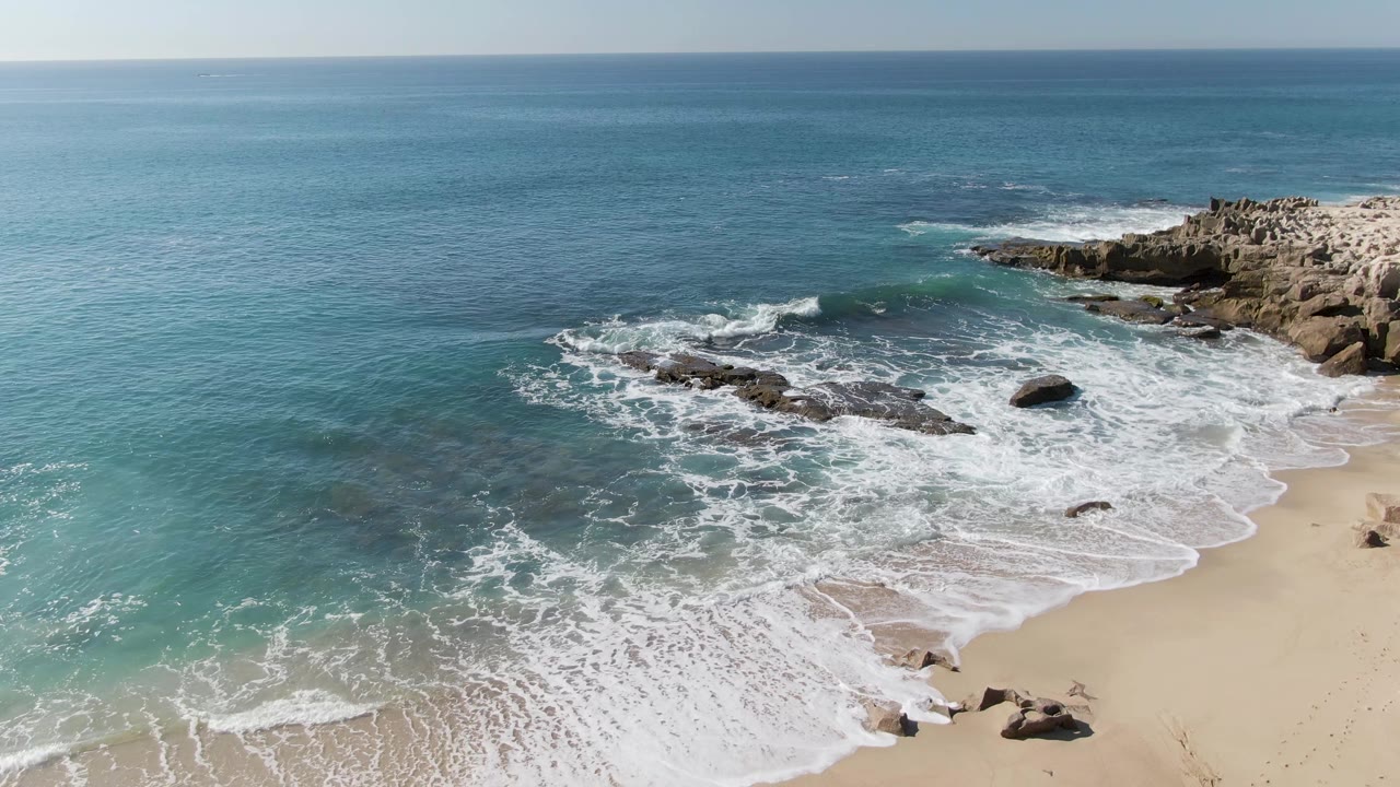 Aerial view at Punta Gorda Beach, Baja California Sur, with turquoise water, white foam around dark rocks, tan sand at lower right, and no visible people.