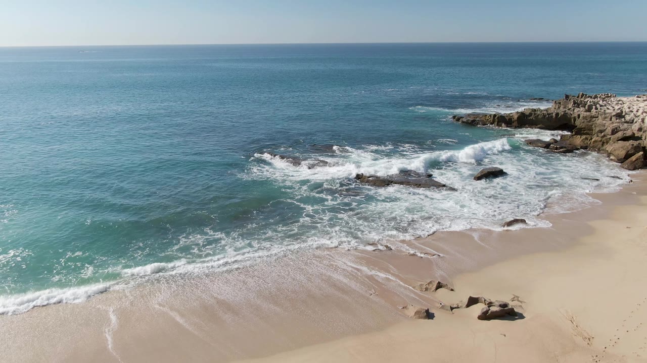 Waves break harder against offshore rocks near a broad sandy beach