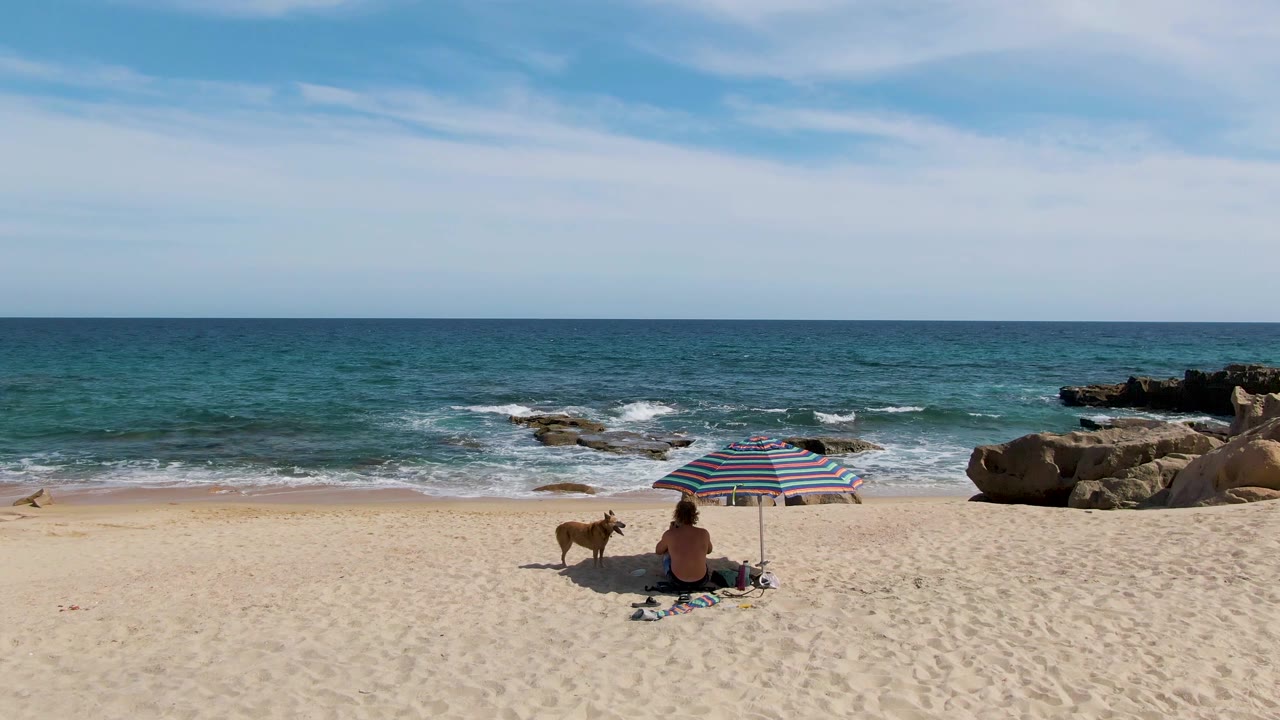 At Punta Gorda Beach in Baja California Sur, a person sits low on the sand under a striped umbrella while a tan dog stands nearby and small waves break beyond the rocks.