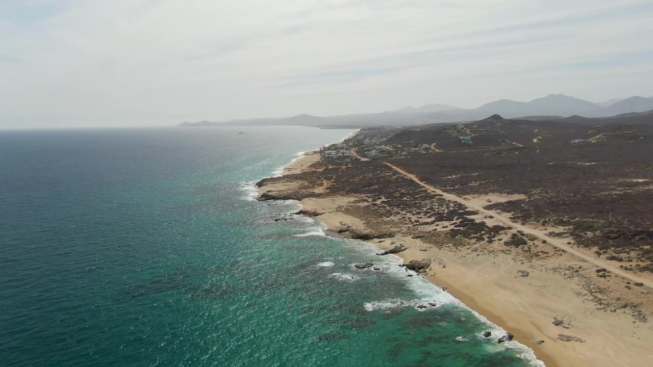 High aerial frame at Punta Gorda Beach showing the shoreline stretching into the distance, a dirt track across scrubland, breaking surf below, mountains on the horizon, and no visible people.