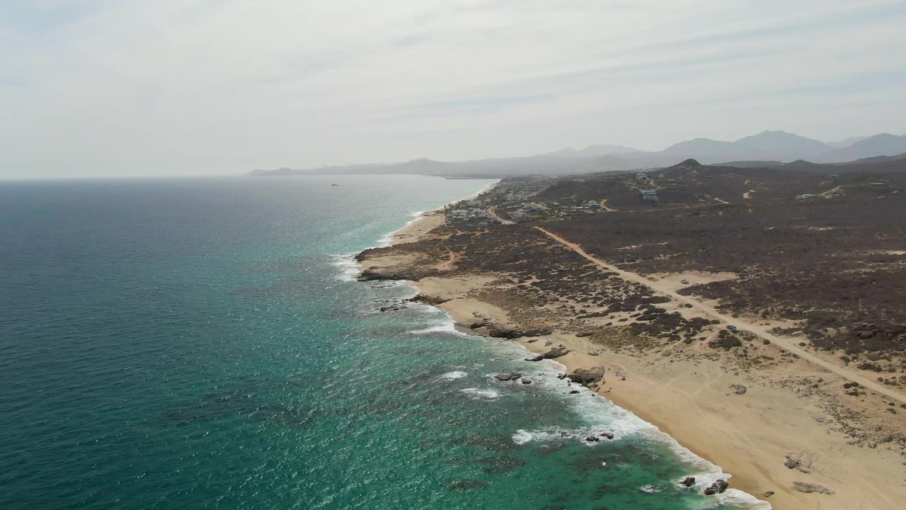 Aerial frame of Punta Gorda Beach with teal water filling the lower left, a band of white surf along the sand, scattered rocks near shore, scrubland inland, and no visible people.