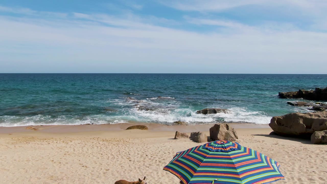 Punta Gorda Beach in Baja California Sur with pale sand in the foreground, blue-green surf around scattered rocks, a striped beach umbrella edge at the bottom, and no visible people.