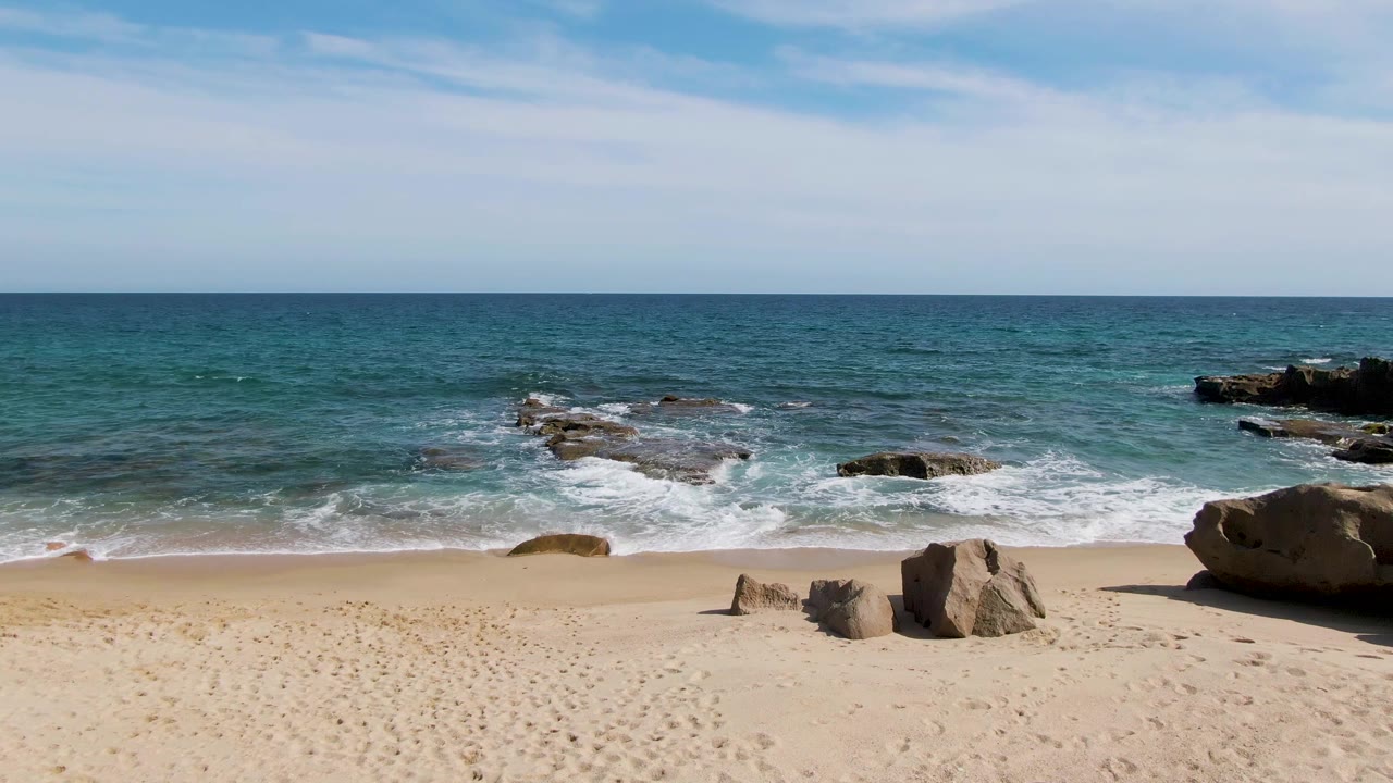 Punta Gorda Beach in Baja California Sur with the surf spreading around a broad flat rock shelf, scattered boulders on the sand, a larger rock at the far right edge, and no visible people.