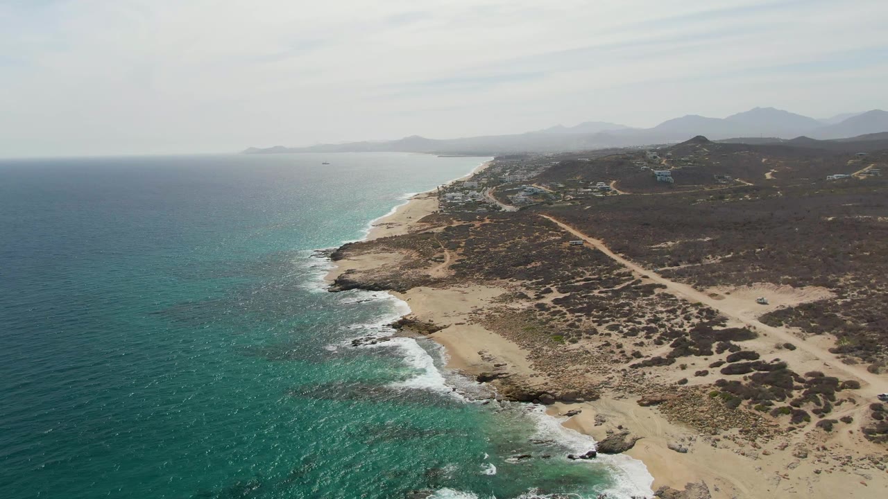 High aerial view at Punta Gorda Beach with teal ocean filling the left side, waves tracing the rocky shore, dry sand and scrub to the right, and no visible people.