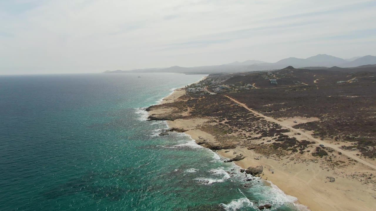 Aerial frame at Punta Gorda Beach showing white surf advancing along the sand, darker rocks near the waterline, sparse dry vegetation inland, a diagonal dirt path, hazy mountains, and no visible people.