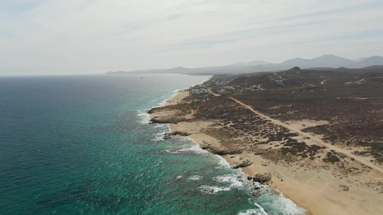 Aerial view of Punta Gorda Beach with deeper blue-green water, scattered white foam patches, a sandy cove edged by rocks, scrub-covered land, faint dirt tracks, distant mountains, and no visible people.