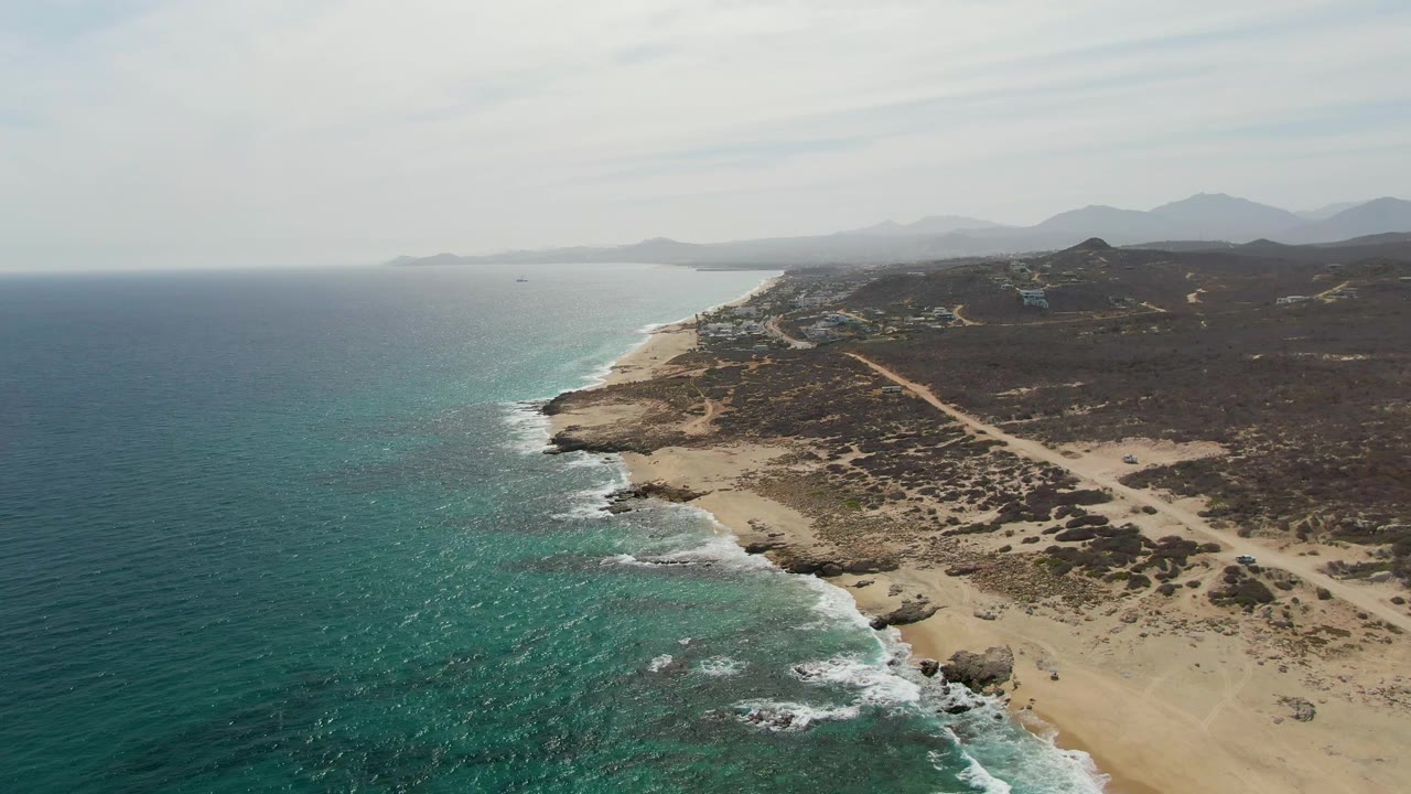 Mid-clip aerial frame at Punta Gorda Beach where the ocean occupies more of the left side, waves meet a narrow sandy beach, rocks break the shoreline, a dirt path crosses the scrubland, and no visible people appear.