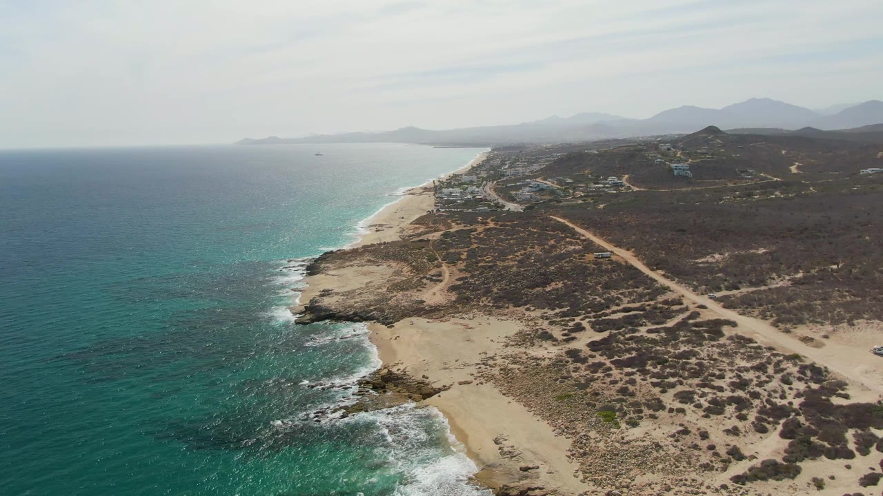 Aerial coastline with turquoise ocean, sandy beach, rocks, scrubland, road, and distant mountains