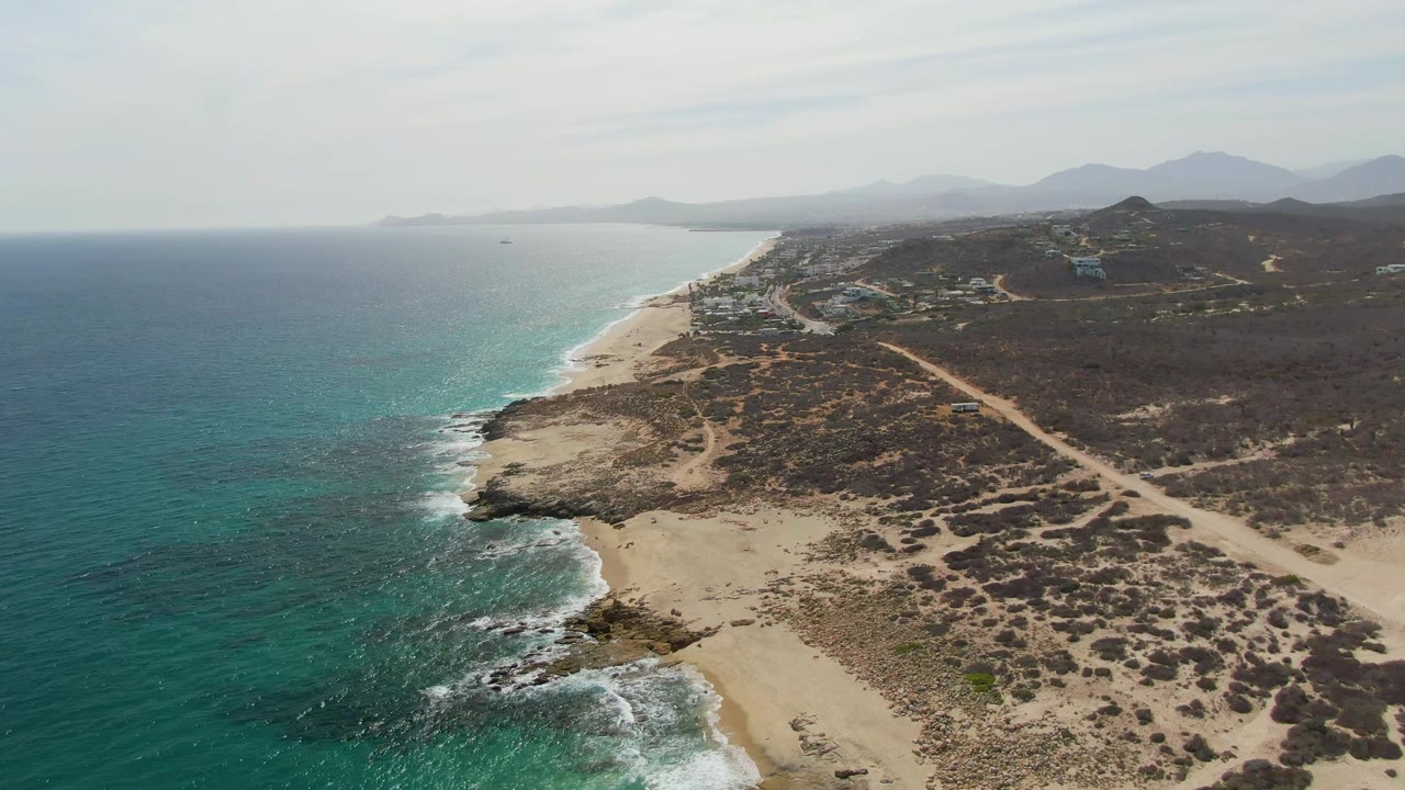 Aerial view at Punta Gorda Beach with the surf line slightly farther along the rocky point, turquoise ocean to the left, pale sand below, dry scrub and a dirt road inland, and no visible people.