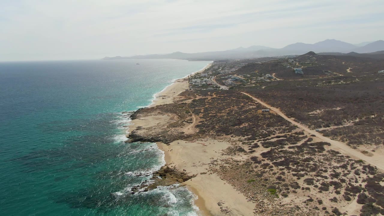 Mid-clip aerial frame at Punta Gorda Beach, showing green-blue water, white foam near shore, sand and rocks in the foreground, a diagonal dirt road through scrubland, distant mountains, and no visible people.