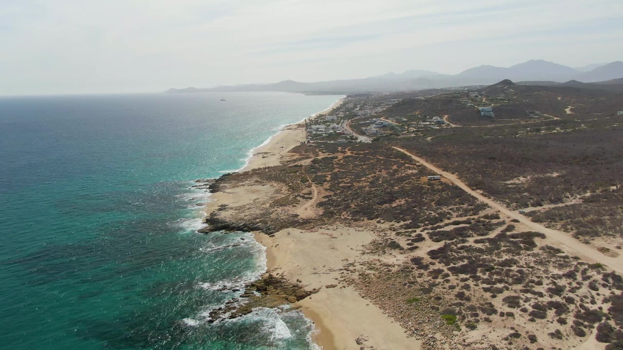 Later aerial frame over Punta Gorda Beach with more foreground beach and rocks visible at the lower edge, turquoise water to the left, dry brush and road inland, hazy mountain ridges, and no visible people.