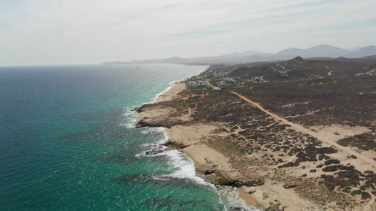 Aerial view of Punta Gorda Beach in Baja California Sur with turquoise water at left, white surf on the sandy and rocky shoreline, dry scrubland, a dirt path inland, distant mountains, and no visible people.