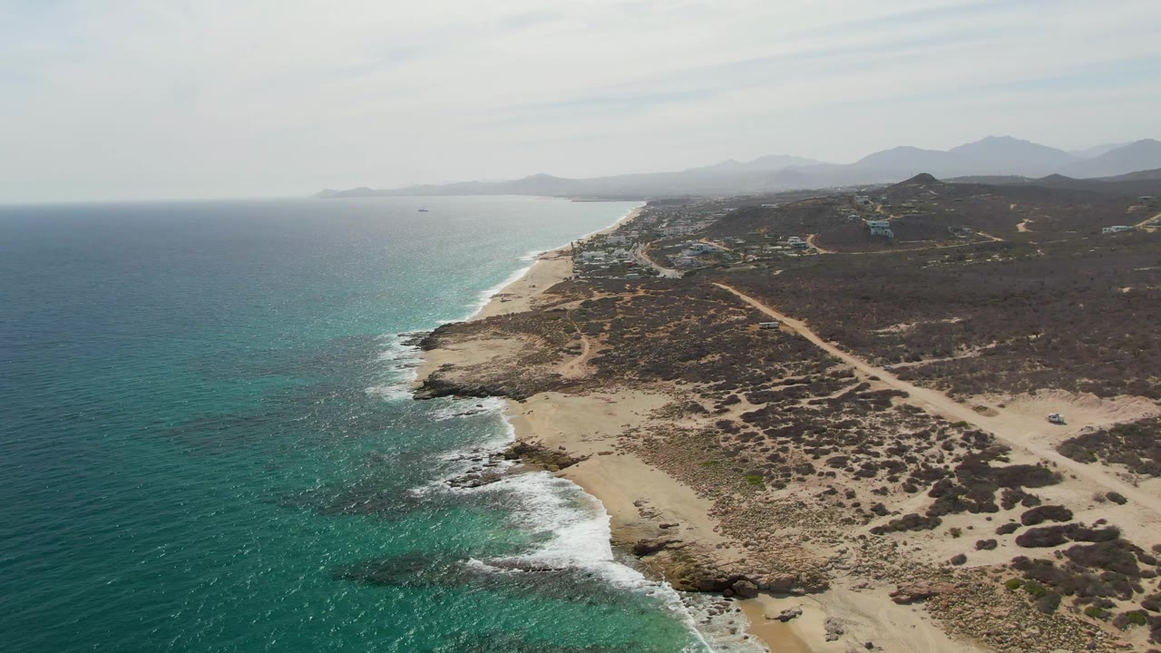 Drone view of waves breaking along a sandy and rocky coastline with scrubland and mountains beyond.