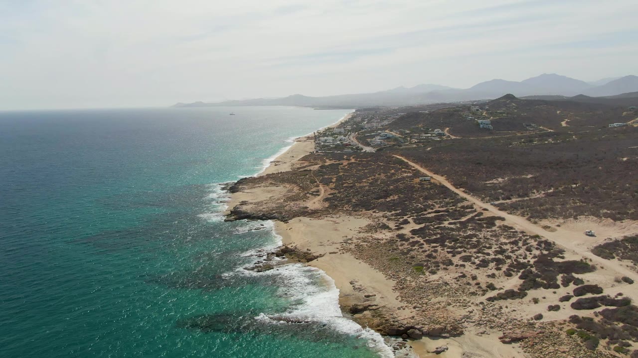 Aerial frame from Punta Gorda Beach showing turquoise ocean and darker reef-like patches near shore, white foam along the coast, a sandy point, dry vegetation, visible dirt routes inland, distant mountains, and no visible people.