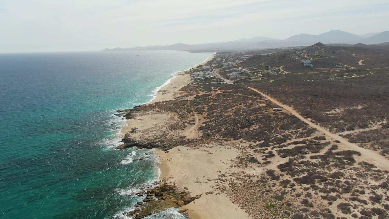 Aerial frame from Punta Gorda Beach in Baja California Sur showing turquoise ocean, a pale sandy beach, rocky shoreline, dry scrub terrain, dirt tracks, distant mountains, and no visible people.