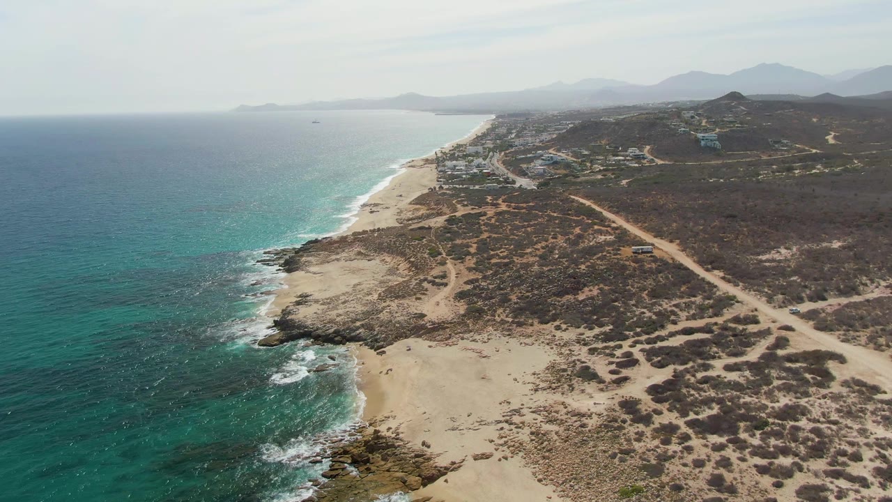 A slightly later aerial view at Punta Gorda Beach shows the rocky point and pale sand below, turquoise water along the left edge, inland dirt roads, scattered coastal structures, distant mountains, and no visible people.