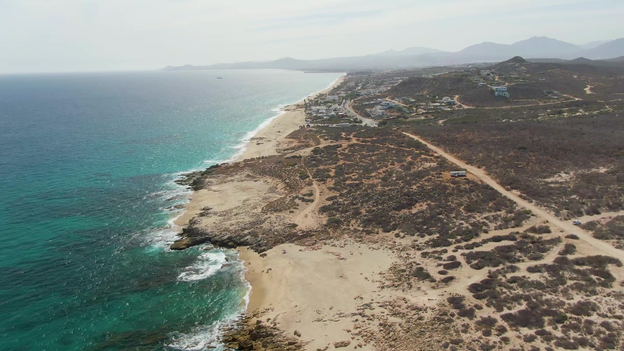 Aerial coastline with waves breaking near rocks, sandy beach, dirt roads, and dry coastal brush