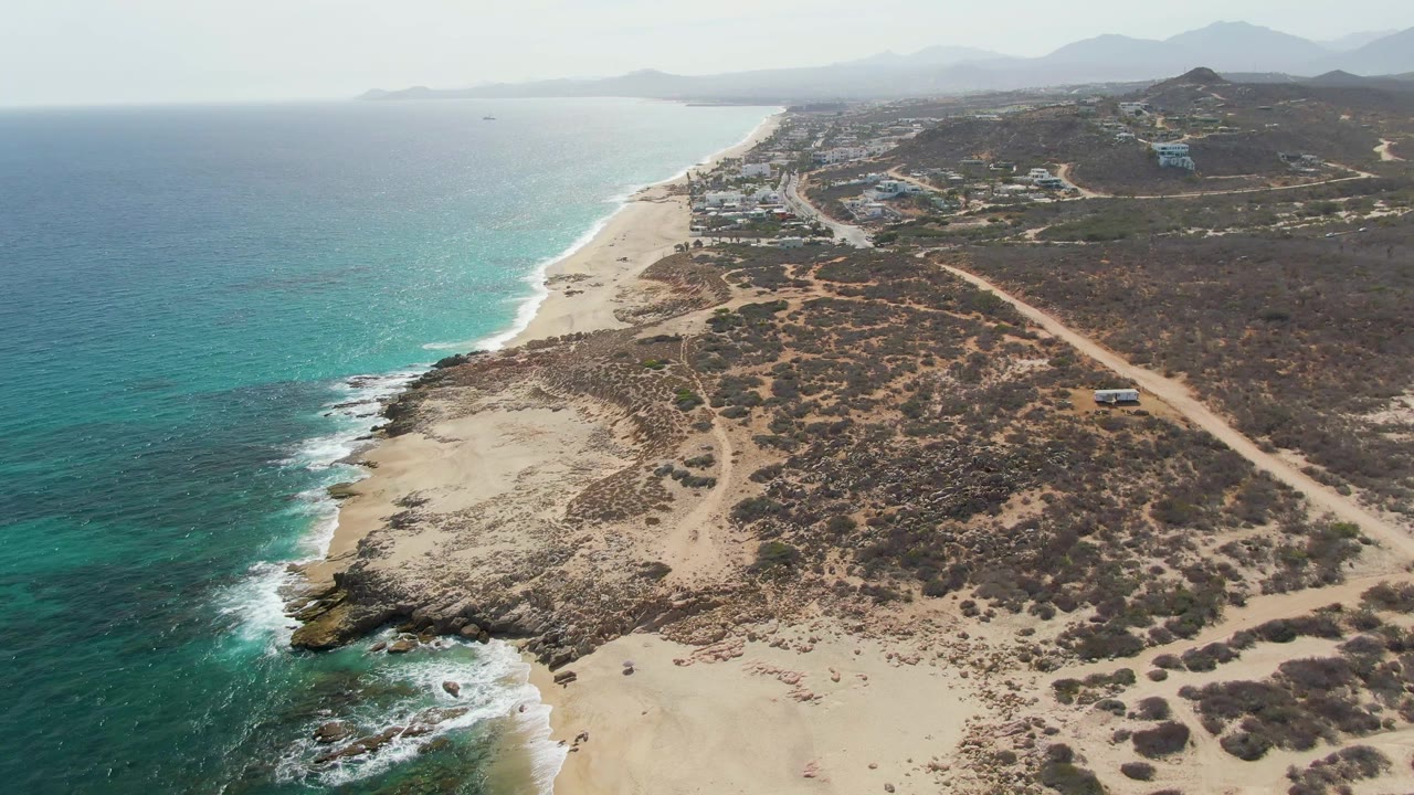 Aerial shoreline with turquoise water, white surf, sandy beach, scrubland, road, and distant mountains.