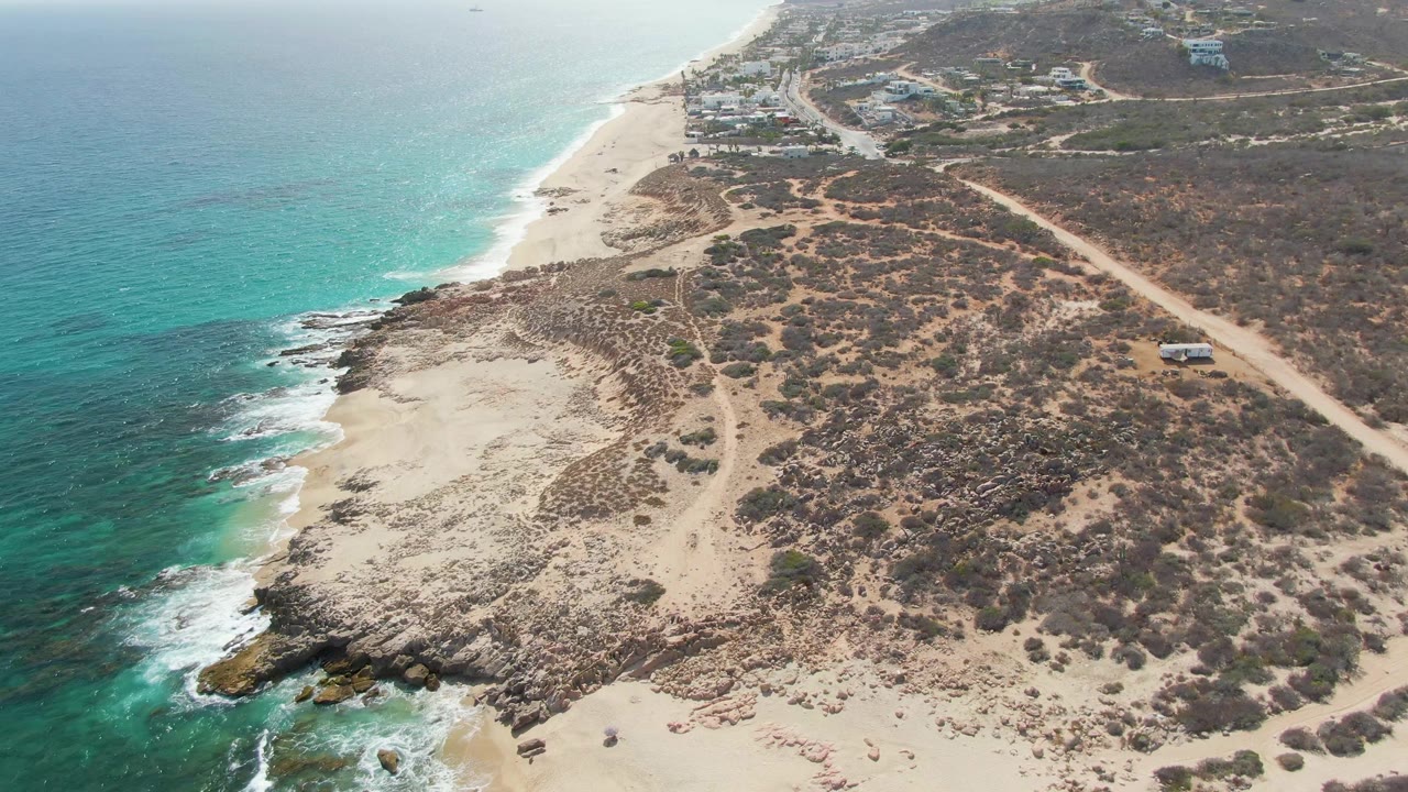 Aerial view at Punta Gorda Beach with foamy waves along the left shoreline, dry scrubland in the center, a dirt road on the right, and a small vehicle visible on the road, with no visible people.