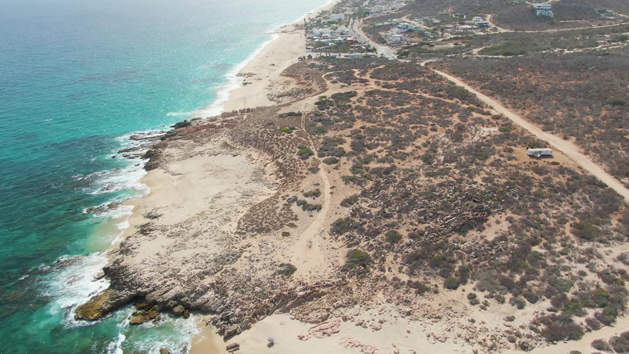 Drone-style frame over Punta Gorda Beach, Baja California Sur, with the coastline running diagonally, deeper turquoise water offshore, scrub-covered sand inland, and the road vehicle farther along the right side, with no visible people.