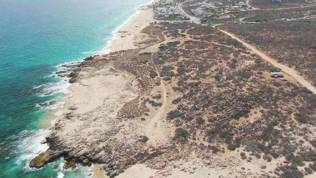Aerial shot of turquoise water washing onto rocky sand beside dry coastal vegetation.