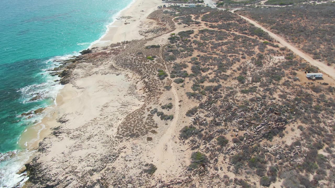 Aerial view over Punta Gorda Beach in Baja California Sur with turquoise water at left, sandy and rocky shoreline below, dry scrubland, a dirt road, a small structure at right, and no visible people.