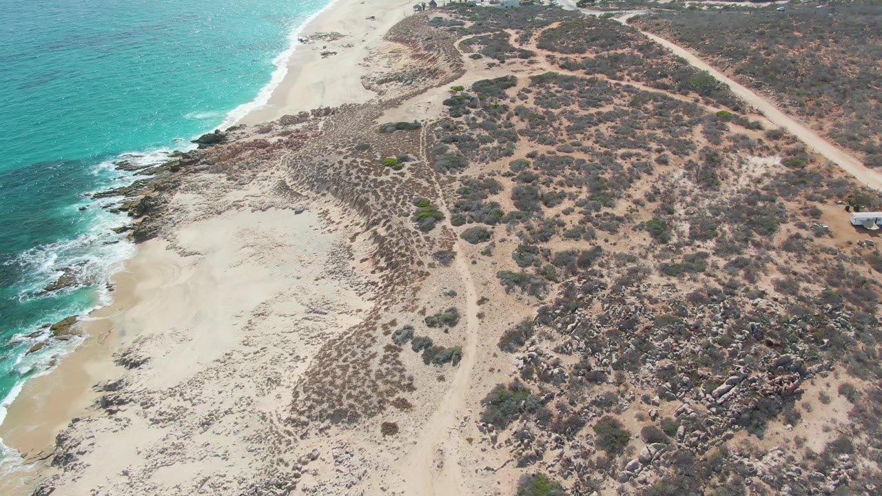 Drone view over Punta Gorda Beach with surf touching the rocky shoreline, a wide sandy patch below, scrubby inland terrain to the right, a dirt road near the top right, and no visible people.