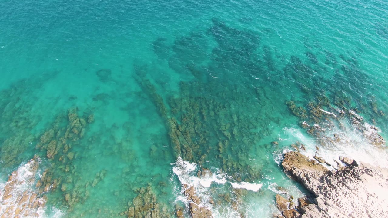 Aerial frame from Punta Gorda Beach, Baja California Sur, with blue-green water revealing long submerged rock shelves while white foam curls around the lower shoreline, with no visible people.