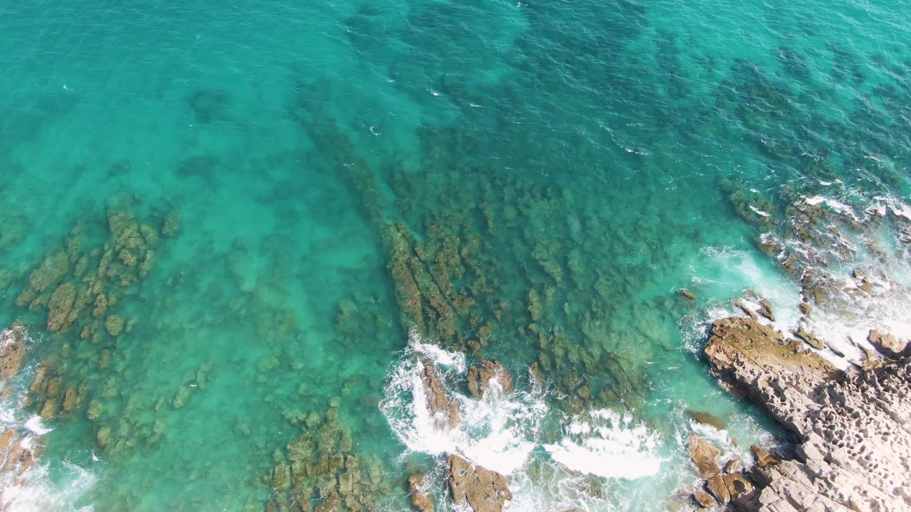 Aerial view over Punta Gorda Beach, Baja California Sur, with transparent turquoise water exposing scattered rock bands and a fresh line of white foam along the lower shore, with no visible people.