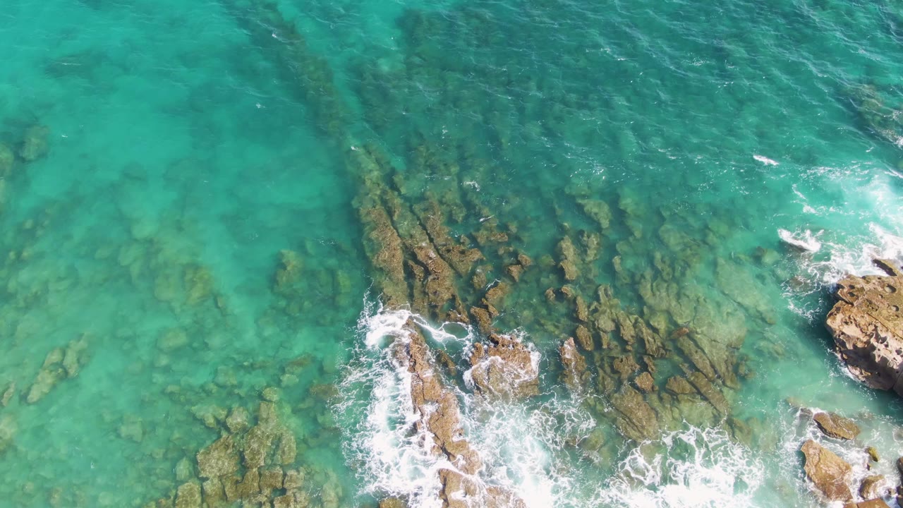 At 0:01, the Punta Gorda Beach aerial view shows rock ridges beneath turquoise water, with surf foam collecting along the right edge and no visible people.