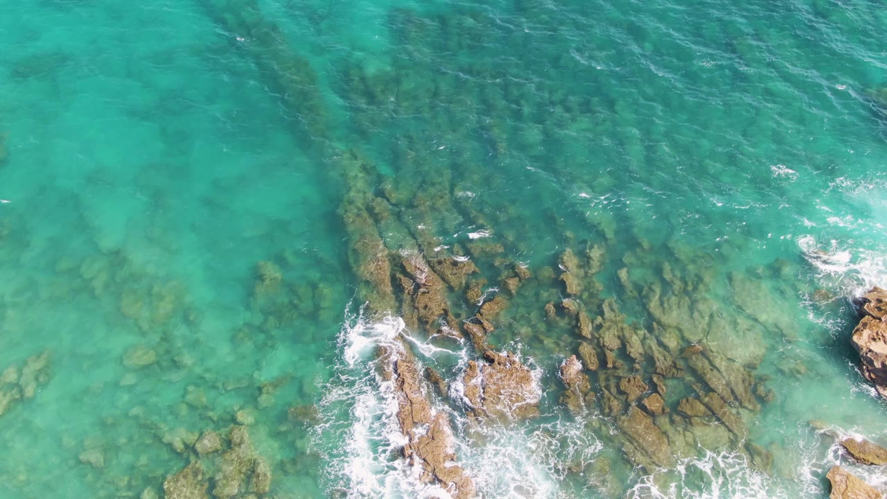 At 0:02, clear blue-green water covers dark rock shelves at Punta Gorda Beach, with fresh white foam around rocks near the lower right and no visible people.