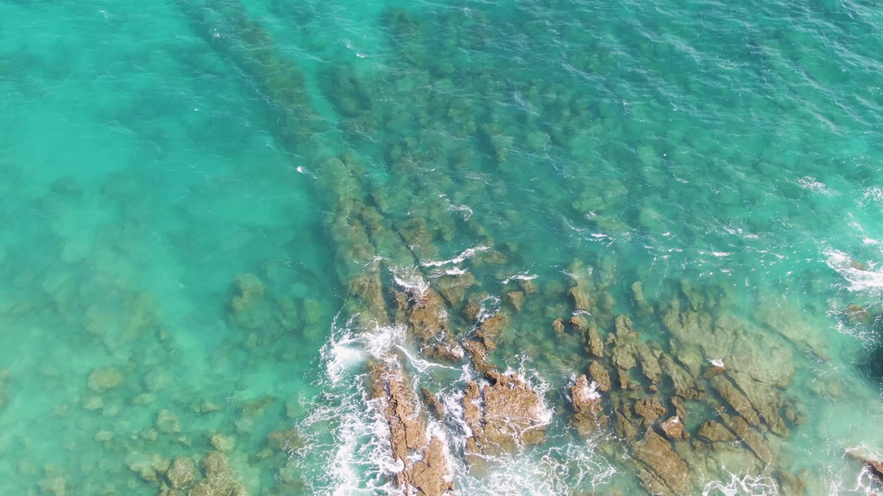 At 0:03, the overhead view at Punta Gorda Beach shows a larger rocky section near the bottom, pale turquoise shallows to the left, and no visible people.