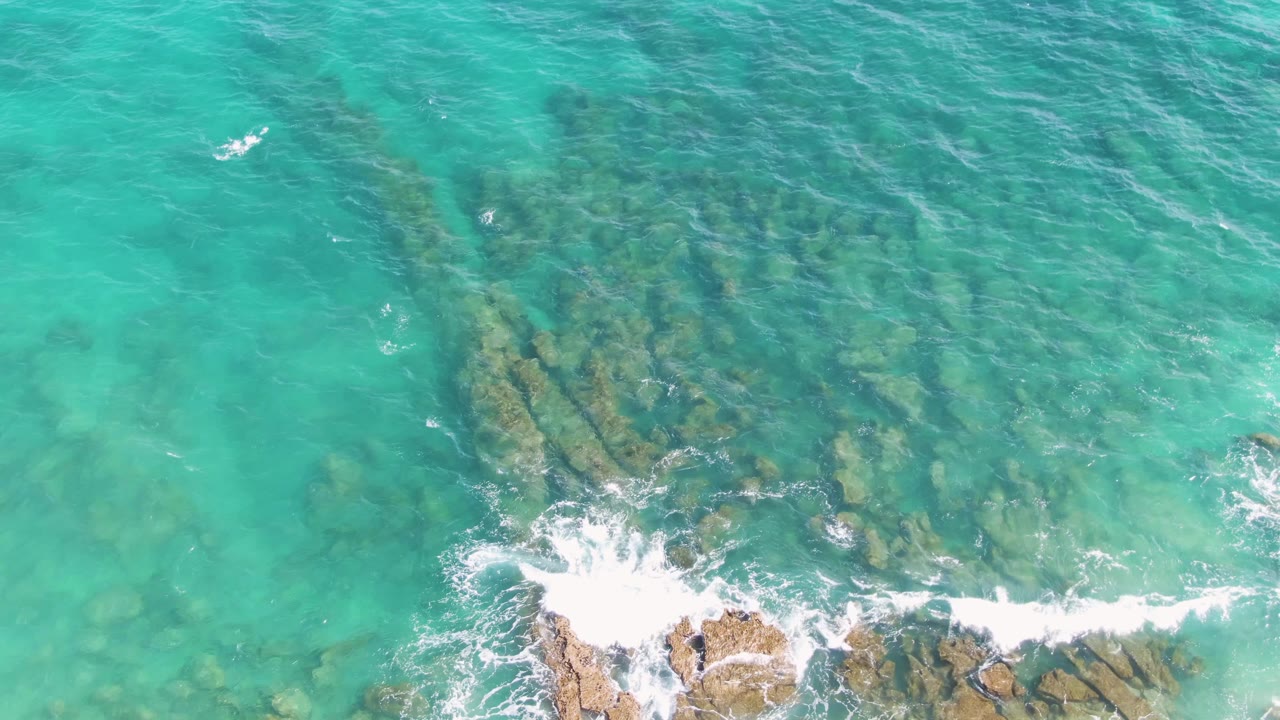 Top-down aerial frame of Punta Gorda Beach turquoise water over submerged rocks, with white foam breaking on the lower-left rocky edge and no visible people.