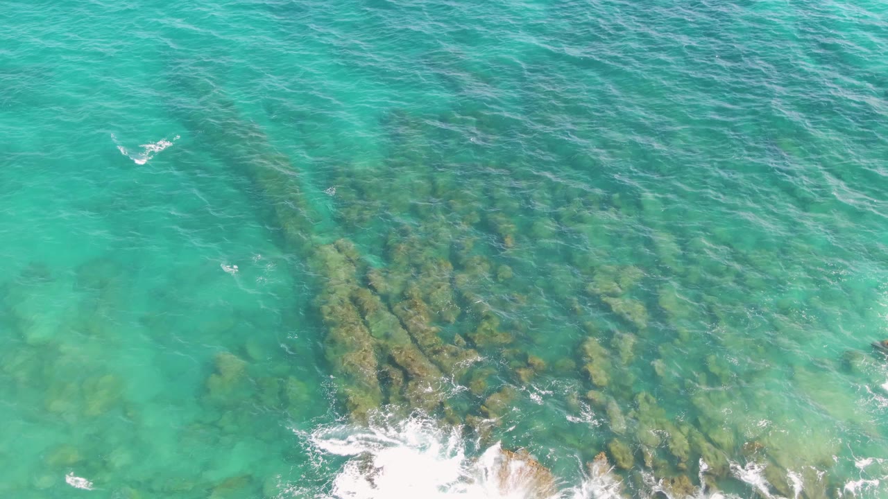 Top-down Punta Gorda Beach frame with turquoise water, darker submerged rocks, and a line of white surf spreading along the lower rocky shore with no visible people.