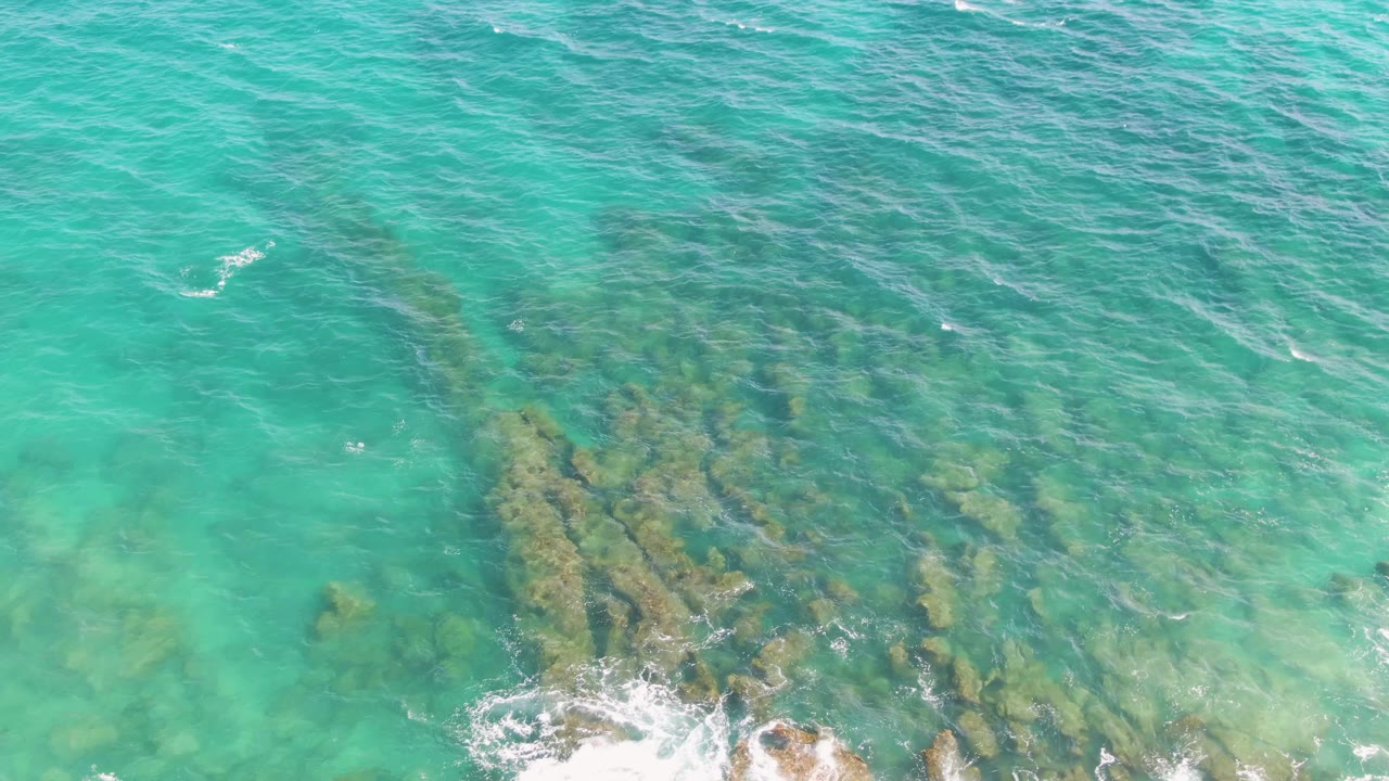 Wide top-down view of turquoise ocean with submerged rocks and small foam patches.