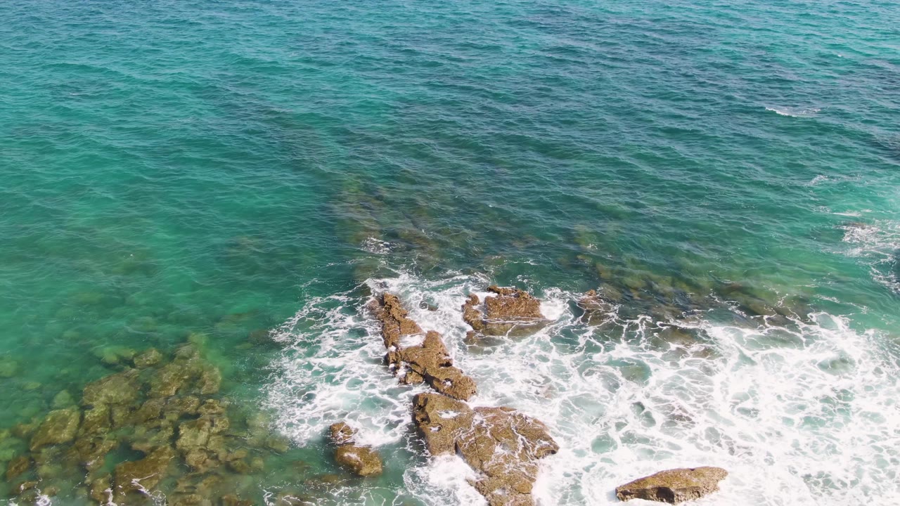 Aerial view of Punta Gorda Beach in Baja California Sur with turquoise water, white foam, and tan rocks near the shoreline, with no visible people in this frame.