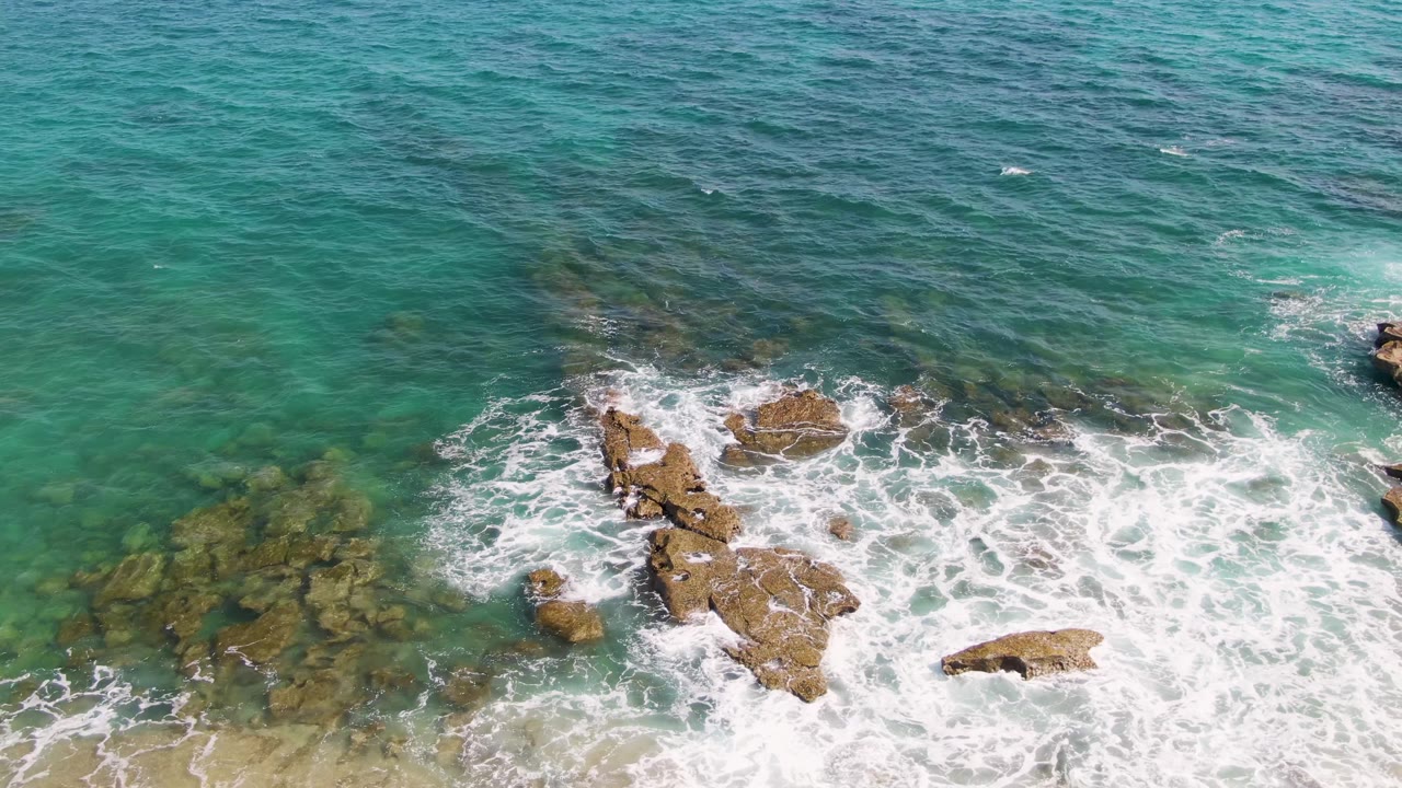 White foam spreads around rocks in clear green-blue water beside sand.