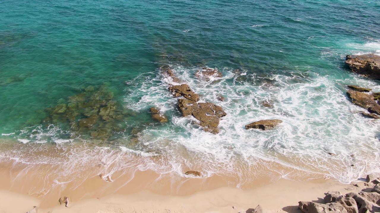 Aerial shoreline view with surf, rocks, sand, and small people on the beach.