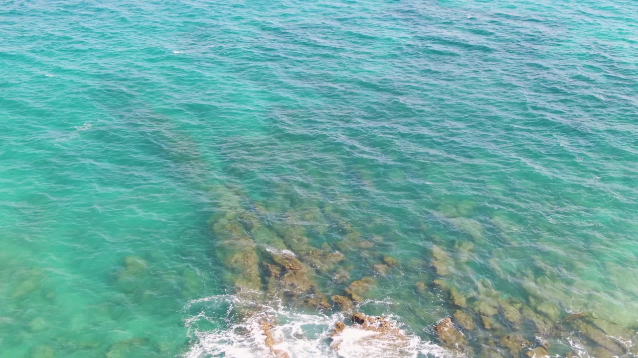 White foam begins to gather on the exposed tips of submerged rocks beneath turquoise water at Punta Gorda Beach, with no visible people.