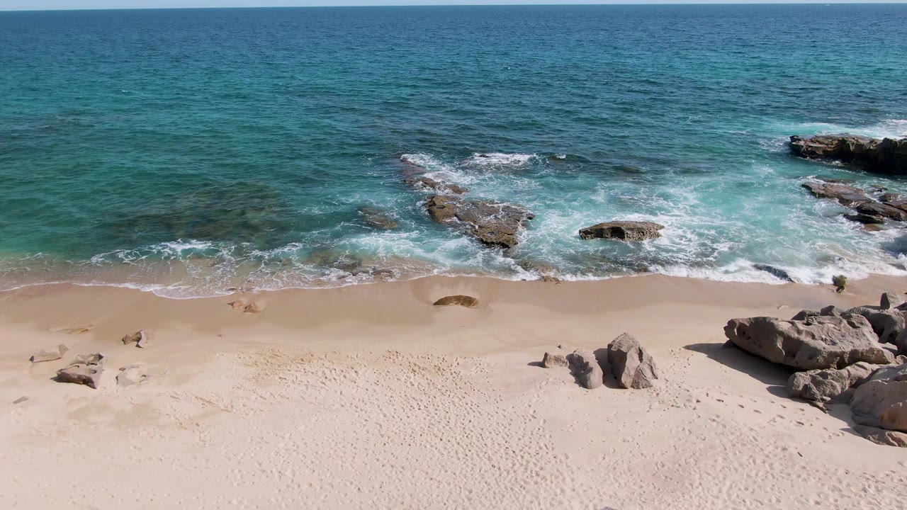 Elevated view of Punta Gorda Beach in Baja California Sur, Mexico, showing pale sand, exposed shoreline rocks, white surf, blue ocean, and no visible people.