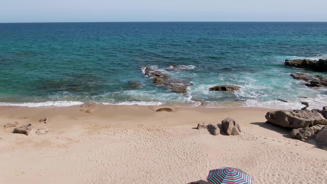 Elevated Punta Gorda Beach view with blue ocean, white surf, scattered rocks, pale sand, and the top of a striped umbrella appearing at the lower edge.