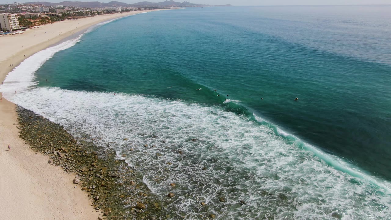 Aerial frame from Punta Gorda Beach, Baja California Sur, showing a broad turquoise wave line beginning to break beside sand and exposed shoreline rocks, with no discernible people visible.
