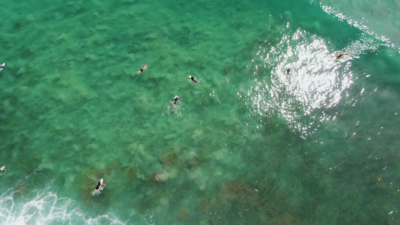 Top-down view of surfers and boards at Punta Gorda Beach, with a band of white foam cutting through the lower part of the turquoise water.