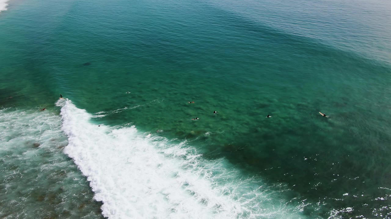 Overhead view at Punta Gorda Beach, Baja California Sur, with a breaking white wave entering from the lower left and small surfers waiting in clear turquoise water.