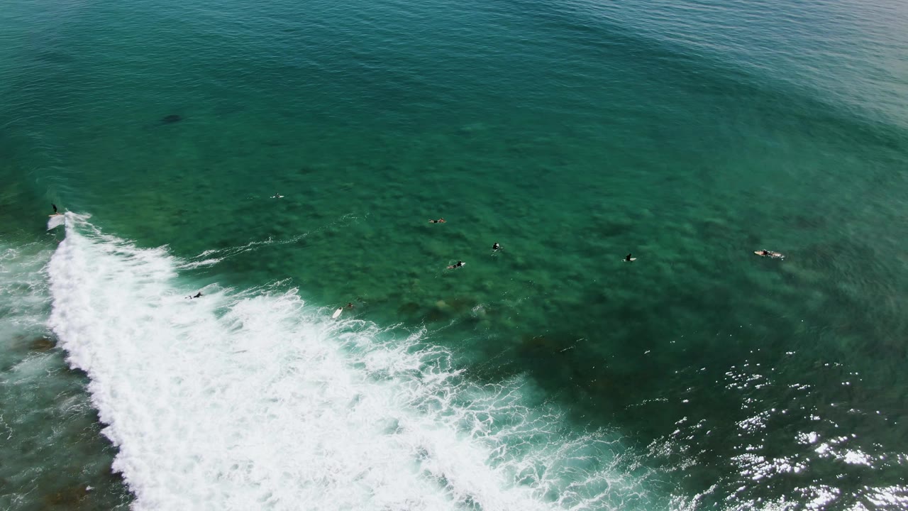 Breaking wave foam crosses clear green water with surfers spaced beyond it.