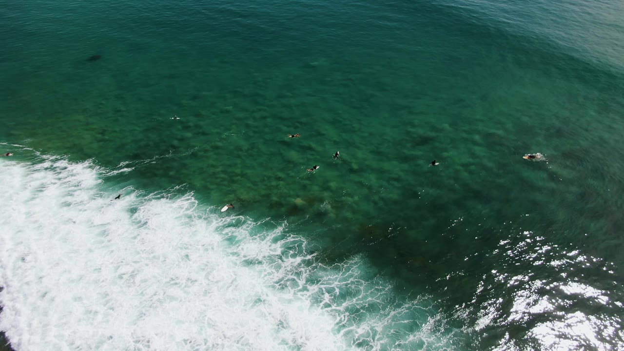 Overhead view of Punta Gorda Beach surf with dense white foam across the lower right, bright surface reflections, and small surfers waiting in the upper water.