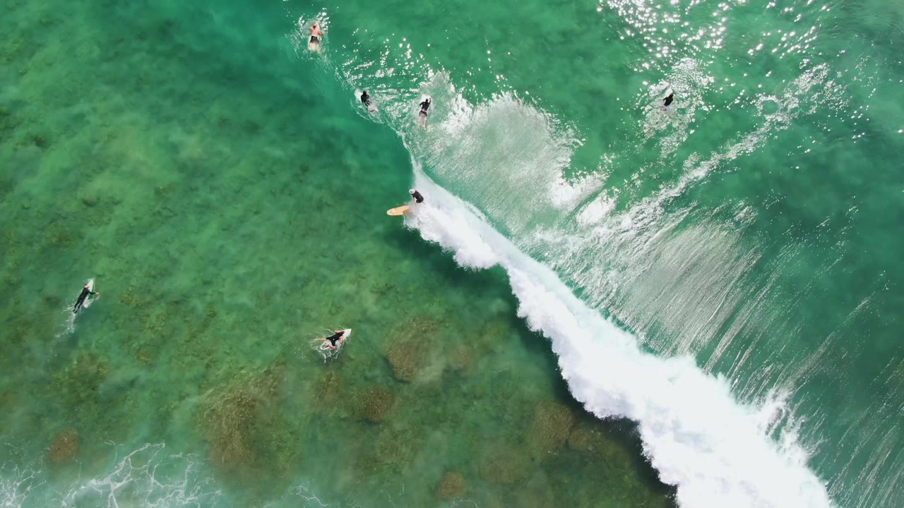 At Punta Gorda Beach, surfers appear as small aerial figures around a stronger diagonal band of whitewater spreading across clear ocean.