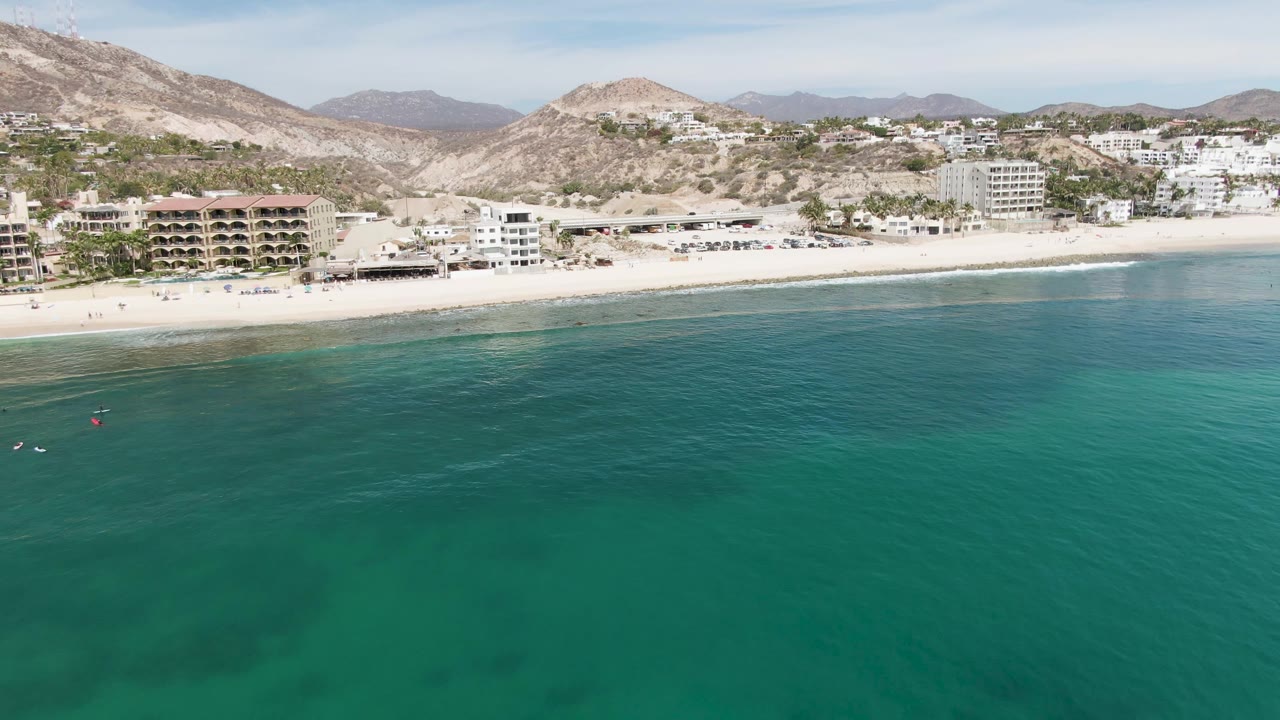 Aerial frame over Punta Gorda Beach in Baja California Sur with turquoise water in the foreground, white sand and coastal buildings across the shore, mountains behind, and tiny distant swimmers visible near the left waterline.