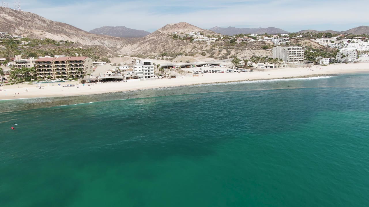 Aerial view of Punta Gorda Beach with clear turquoise water dominating the lower frame, a pale sand shoreline, beachfront buildings, dry mountains, and a few tiny distant swimmers near the left side.